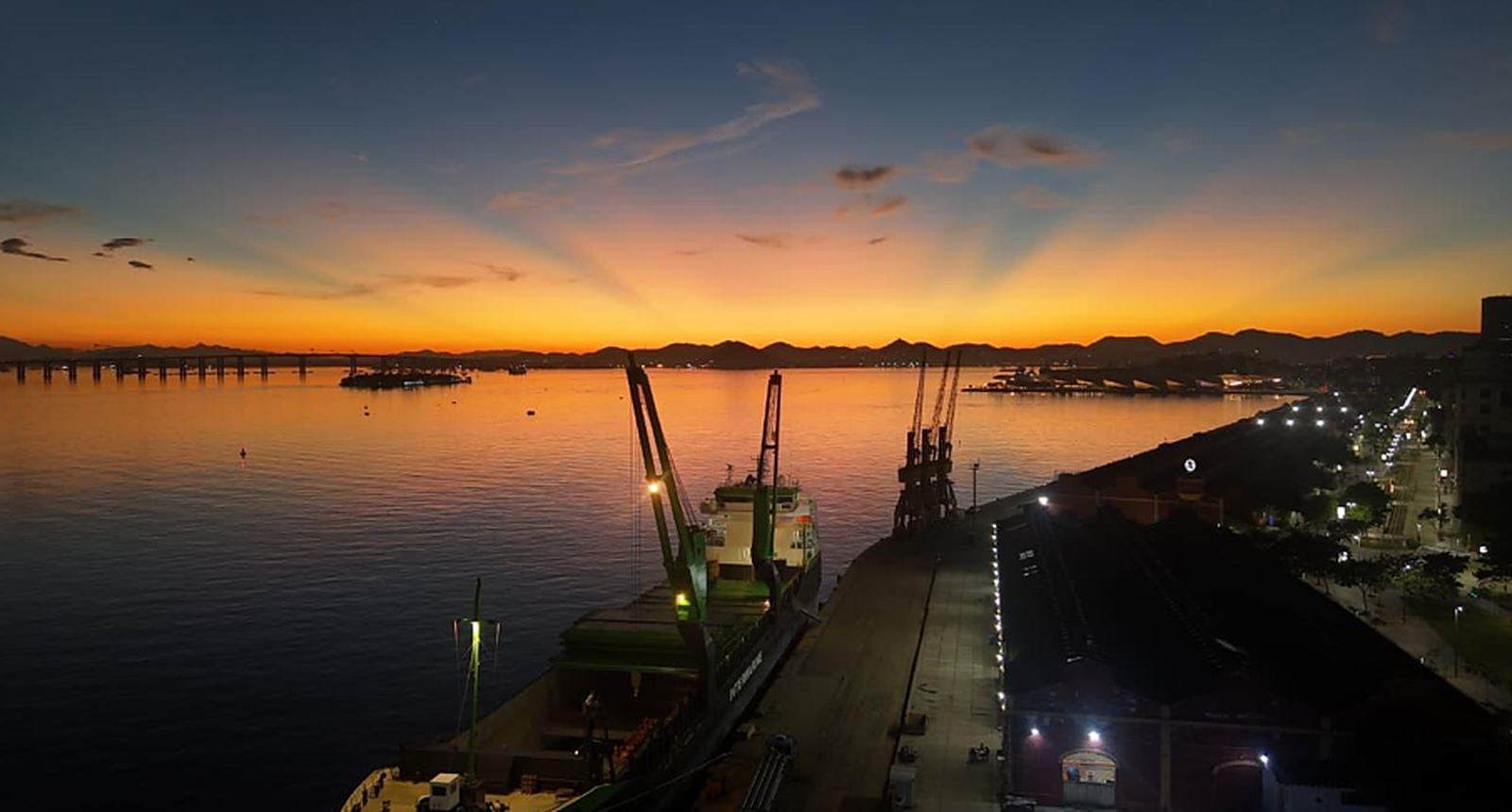 Cais do Porto do Rio de Janeiro ao pôr do sol, com navio atracado, guindastes e a Ponte Rio–Niterói ao fundo
